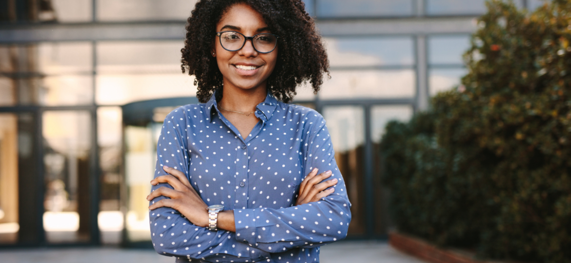 Retaining Talent Graphic: Photo of a young woman in a blue button up. Her arms are crossed and she is standing confidently in front of a glass building. She's smiling.