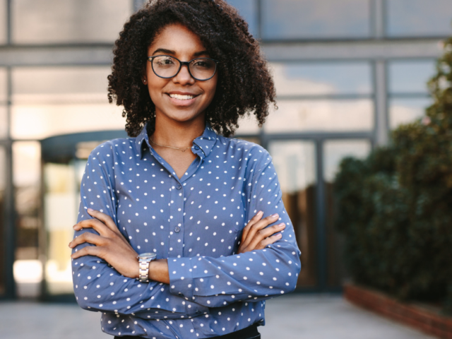 Retaining Talent Graphic: Photo of a young woman in a blue button up. Her arms are crossed and she is standing confidently in front of a glass building. She's smiling.
