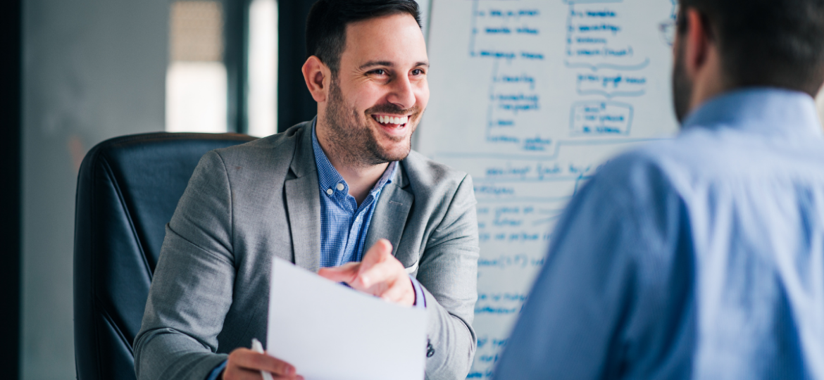 How to Work with a Recruiter to Find a Job Blog Graphic - Image showcases a man in a blazer holding a resume while talking to someone off camera.