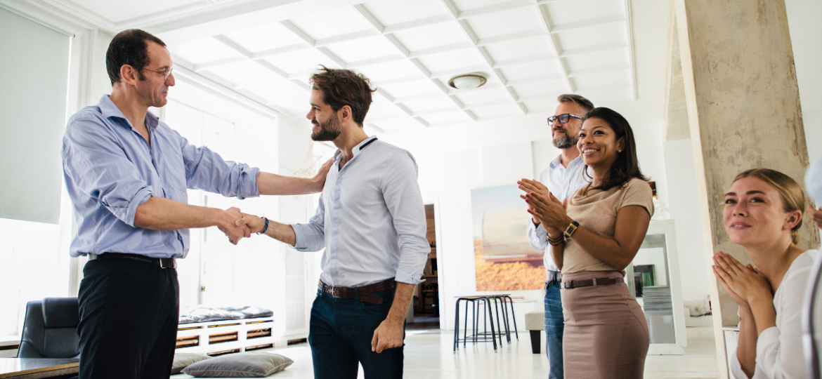 How to Ask for a Promotion Blog Graphic that showcases two men in button ups shaking hands while other coworkers are clapping.